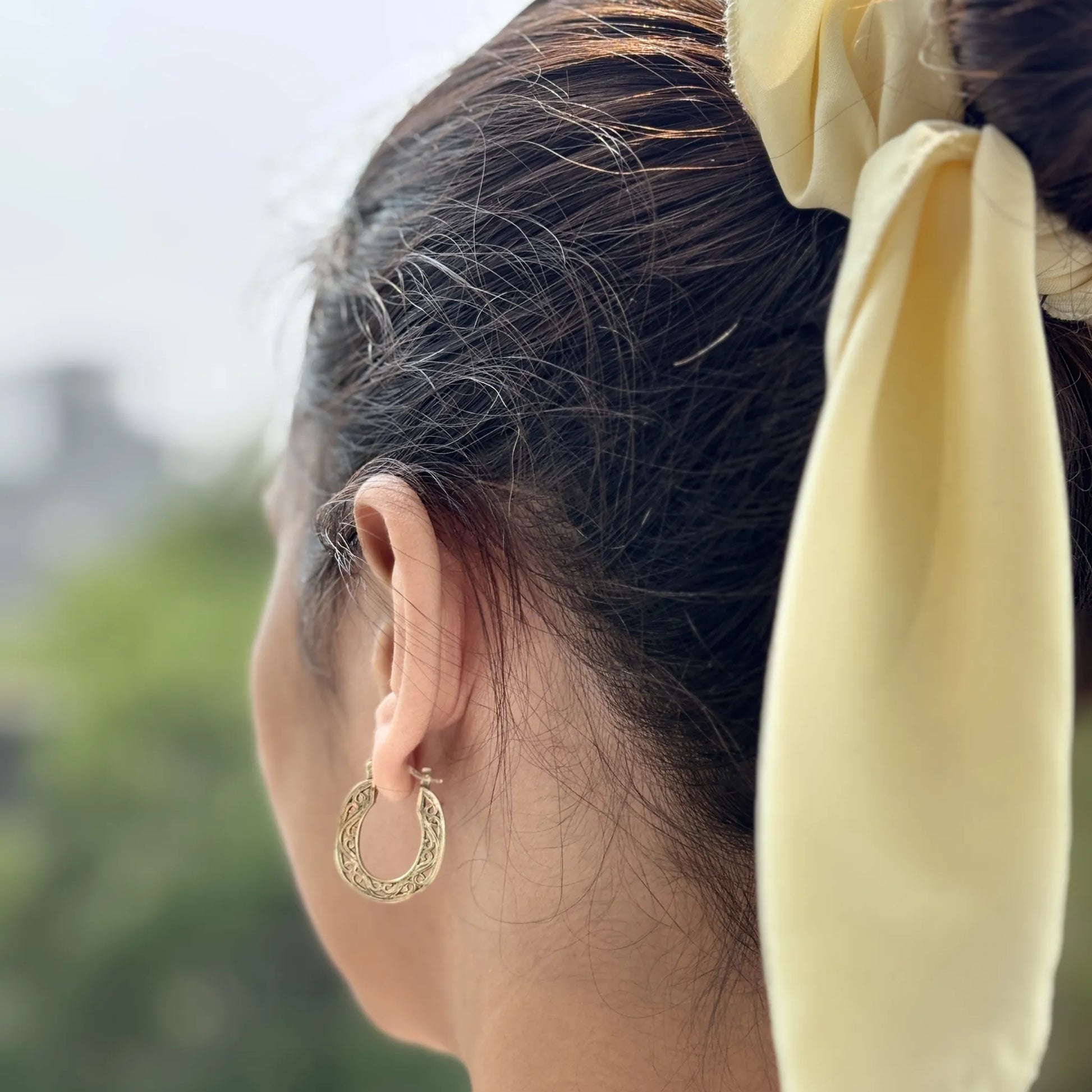 Heritage-style brass hoops worn on ear, hair tied with yellow ribbon