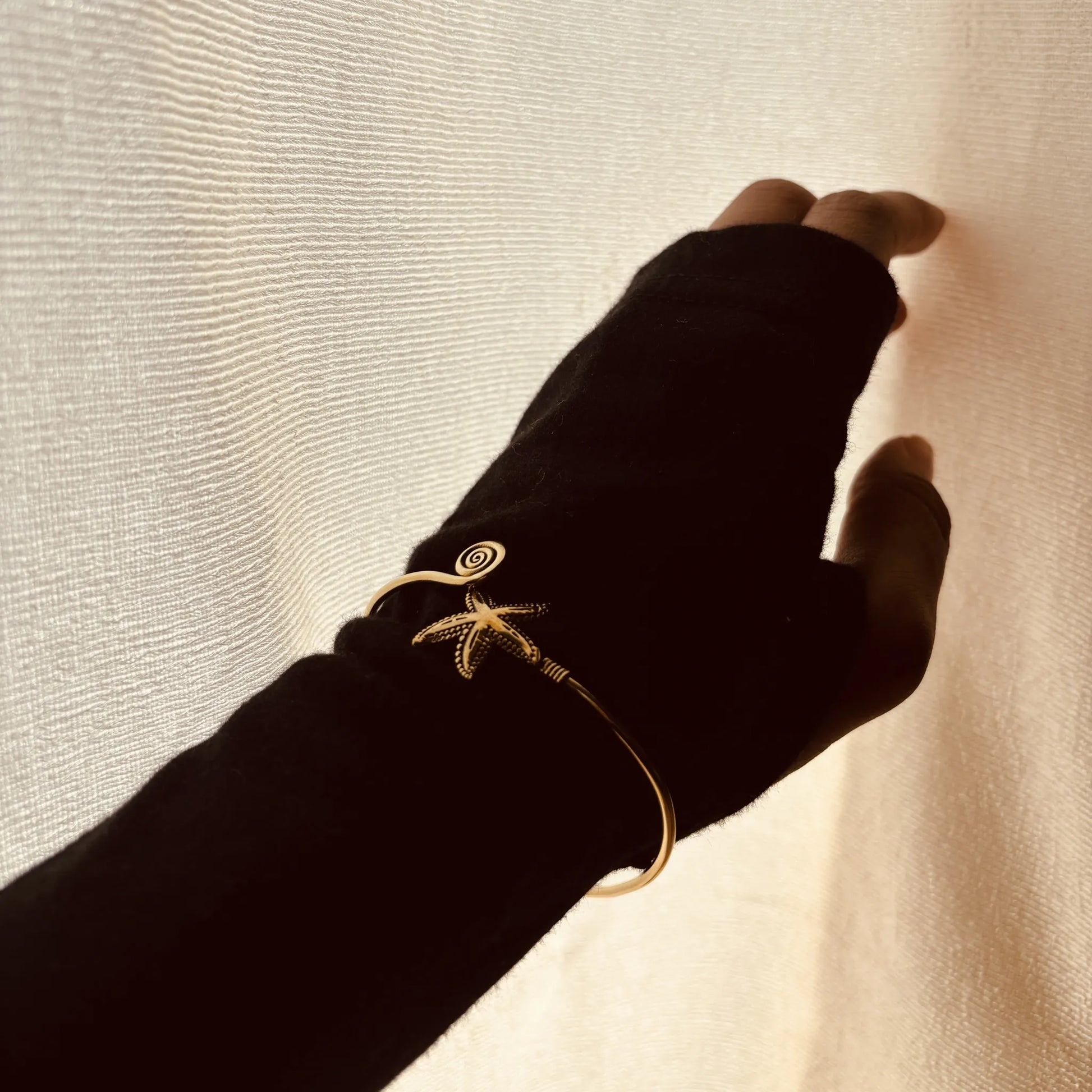 Brass spiral and starfish cuff on wrist reaching toward light through white curtain